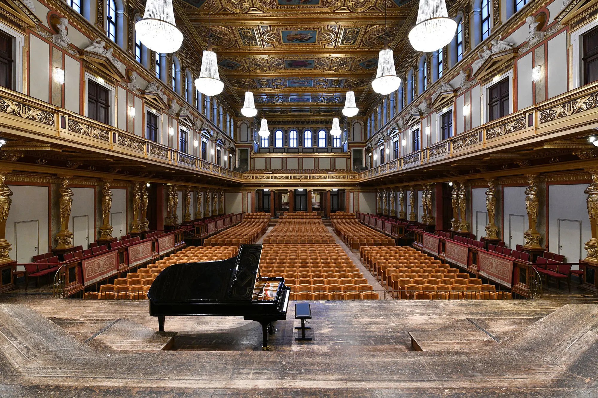 Grand piano on stage facing rows of empty seats in the Musikverein in Vienna with chandeliers and detailed ceiling designs