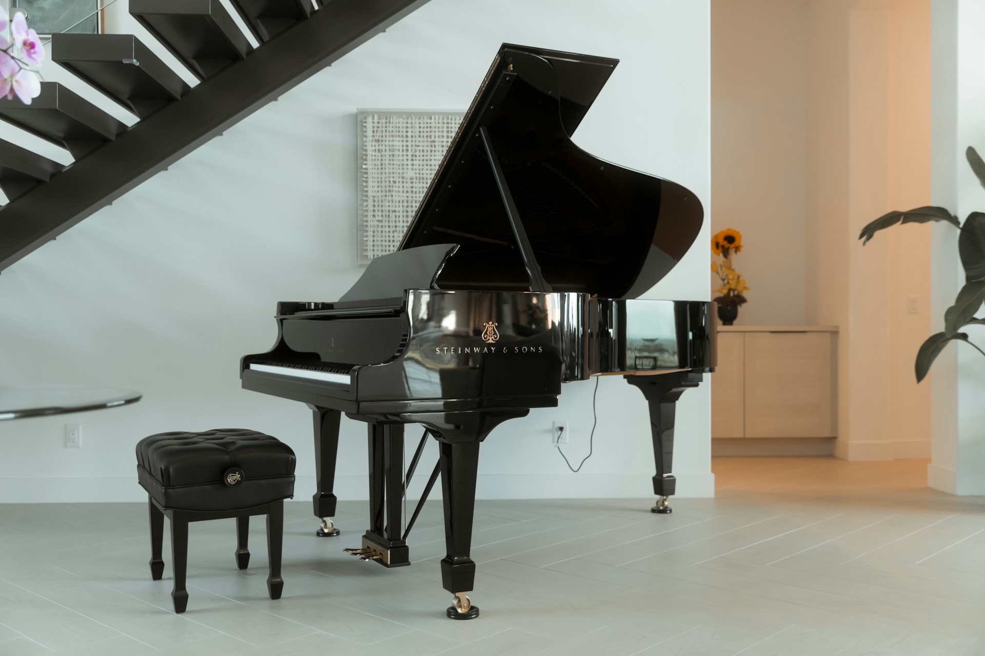 Black grand piano with bench in a modern room beneath a staircase, with white walls and a plant in the background