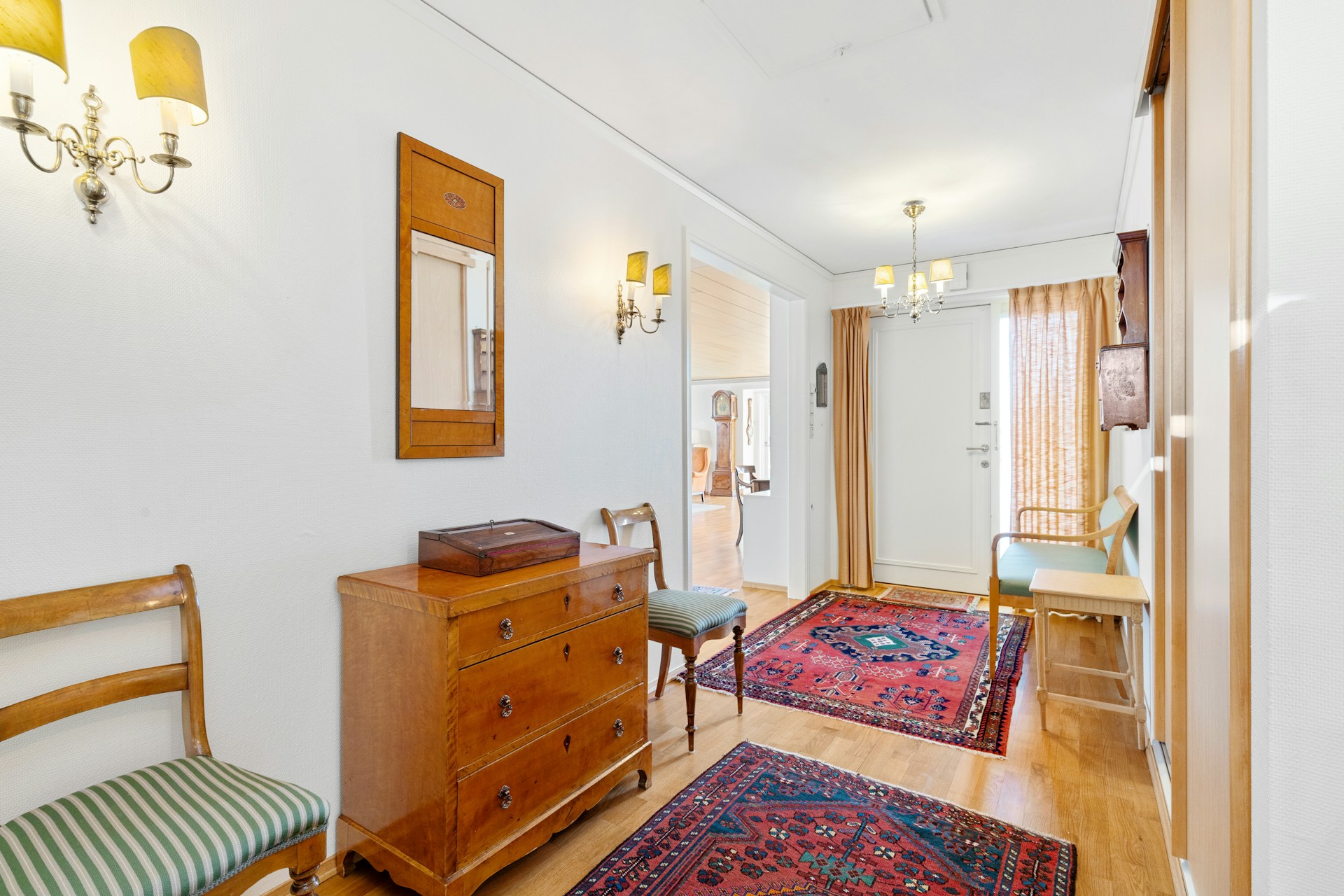 Bright hallway with wooden furniture, chairs, wall lamps, and patterned rugs leading to a door and adjacent room