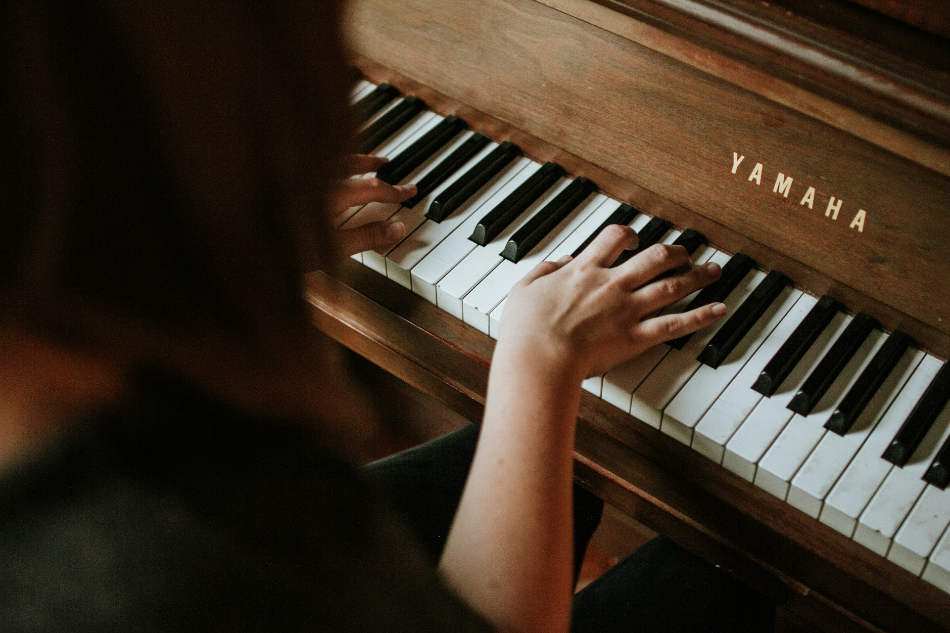 Hands playing a Yamaha piano keyboard with wooden casing, viewed from above with part of a person visible on the left