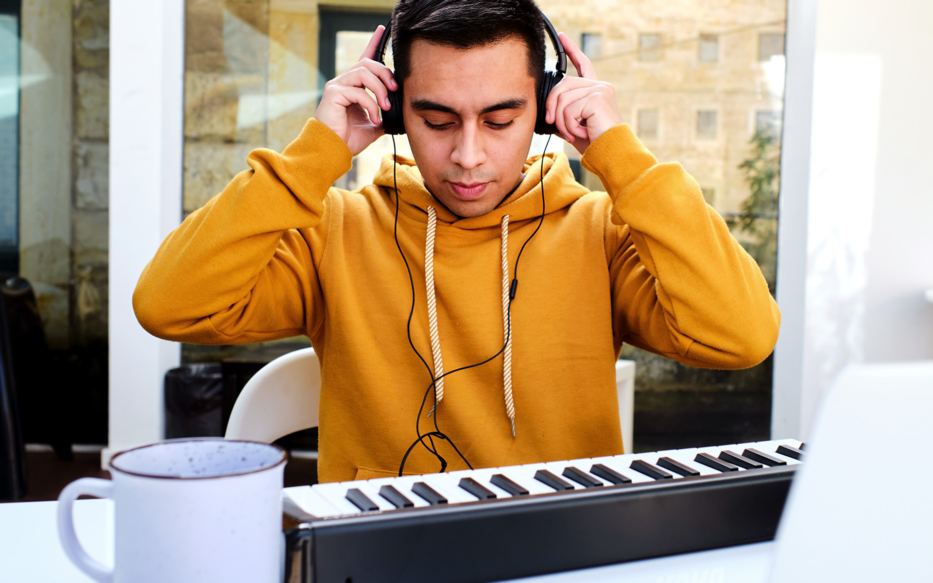Person in yellow hoodie wearing headphones adjusting them while sitting at a keyboard with a mug on the table nearby
