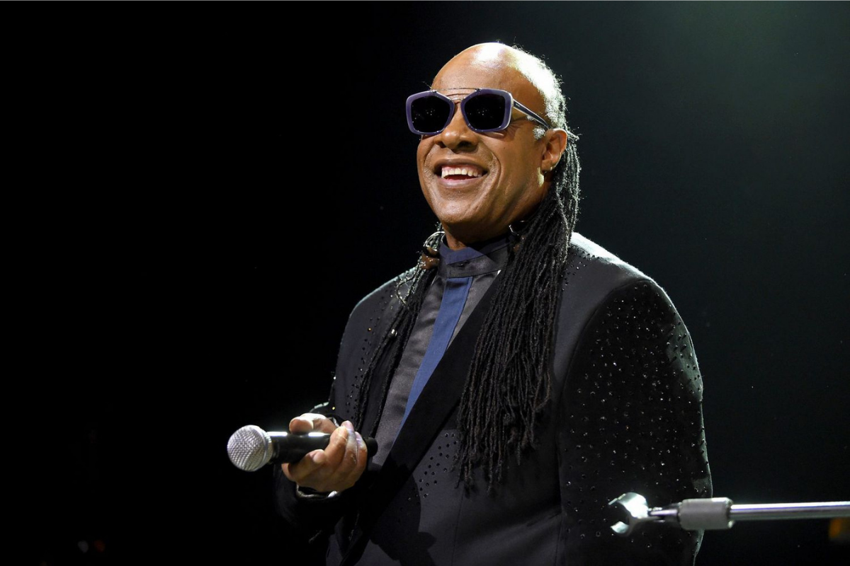 Smiling performer wearing sunglasses holds microphone onstage under spotlight