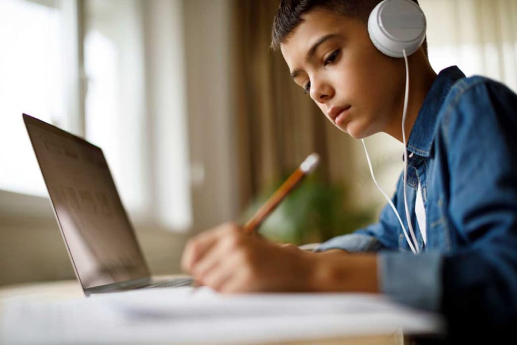 Teen student wearing headphones studies at a laptop while writing notes