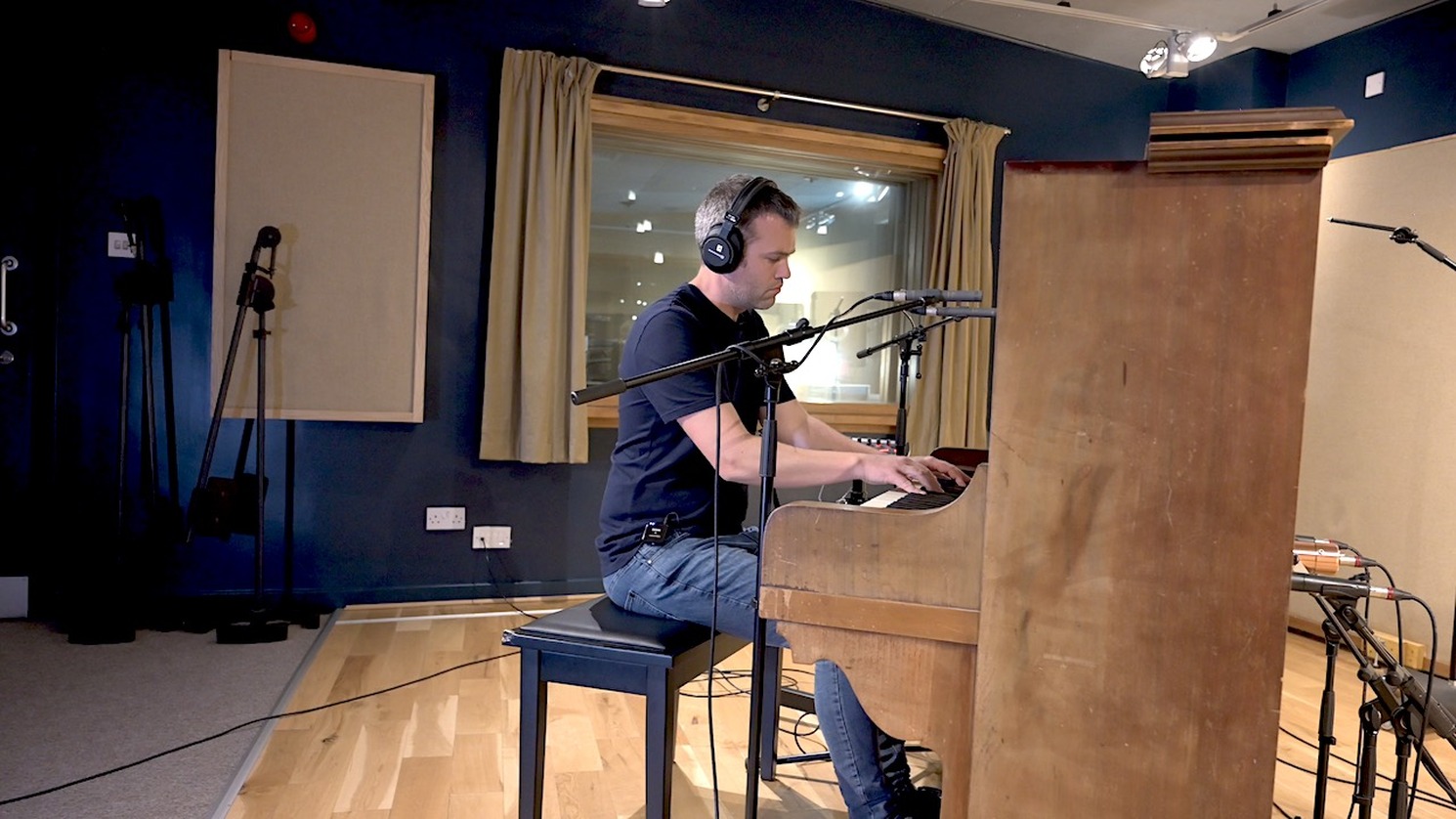 Man wearing headphones playing an upright piano in a recording studio with multiple microphones