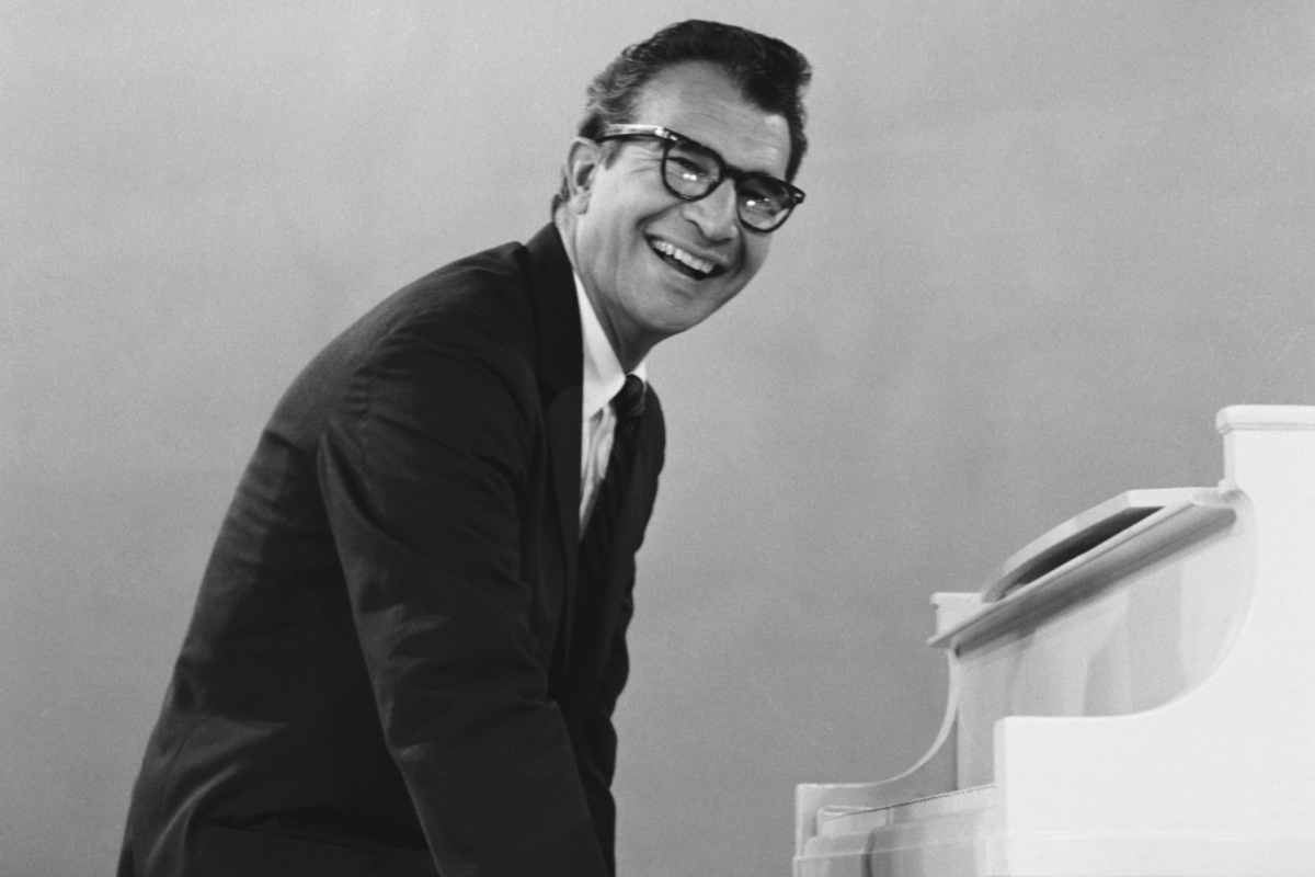 Smiling man wearing glasses standing beside white piano
