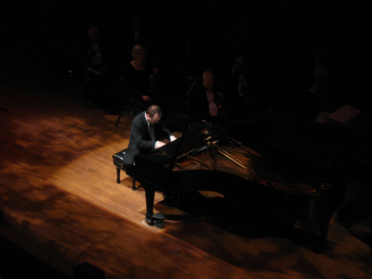 Pianist performing on grand piano on stage, spotlight illuminating musician with audience and orchestra in dark background