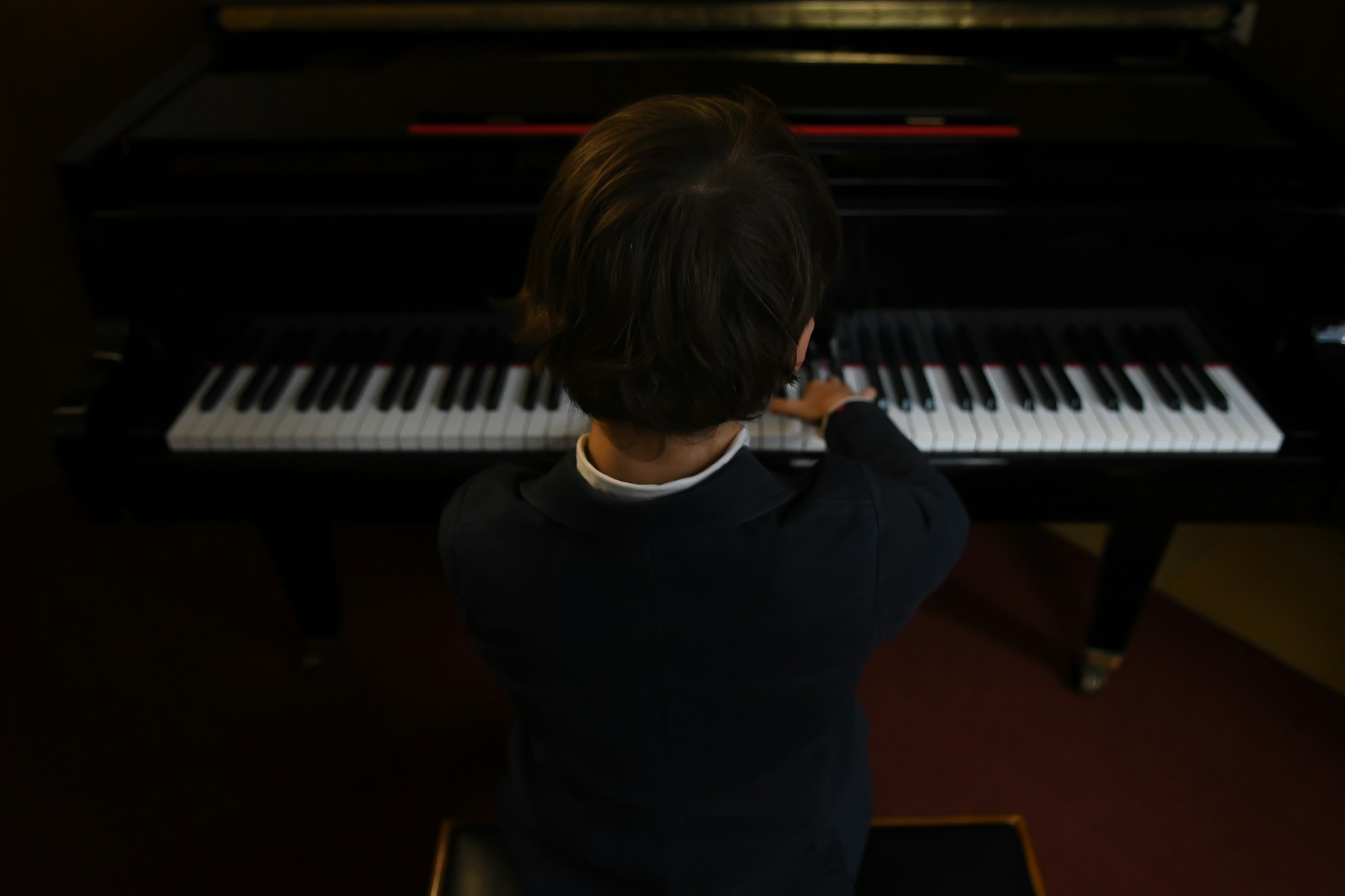 Young child sitting at a piano playing keys viewed from behind