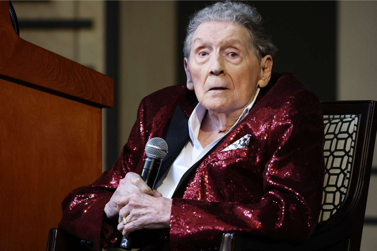 Elderly person in sequined jacket holding microphone while seated