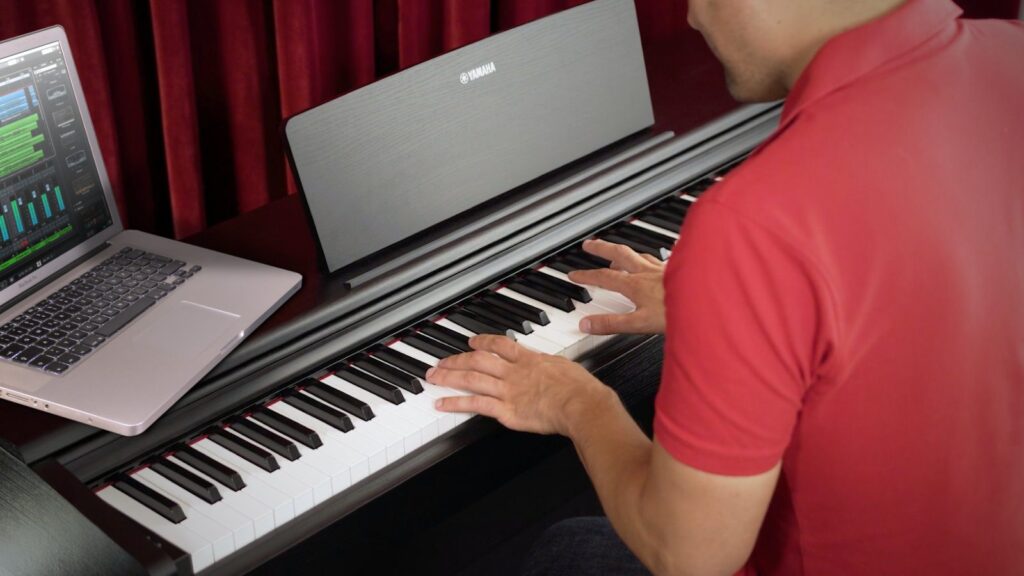 Person playing a digital piano connected to a laptop, viewed from behind in a home studio setting